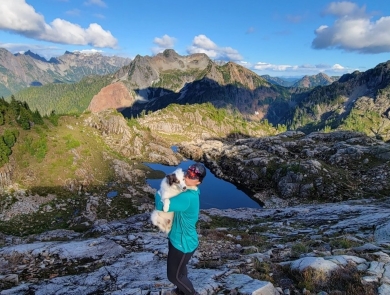 A woman holds a white dog with majestic mountains in the background