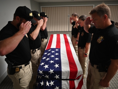 The honor guard stands over a coffin draped in an American flag. Each of them is saluting and looking down at the coffin/flag.