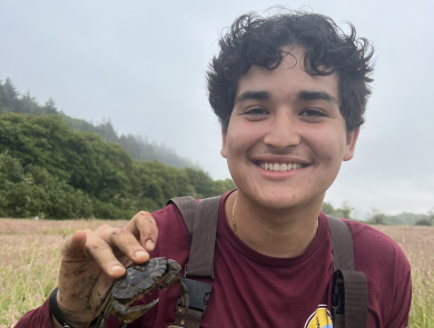 AmeriCorps service member in waders holding a European green crab. Marsh vegetation, trees, and overcast sky in the background.