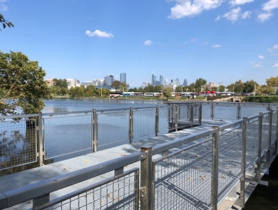 View of a pier with metal railings and synthetic decking jutting out into a river, with a city skyline in the distance