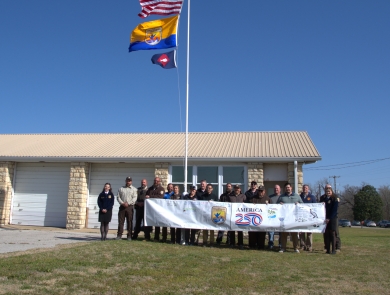 People hold FWS A250 banner at base of flagpole with US flag, FWS flag, fish pennant flying