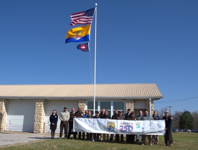 People hold FWS A250 banner at base of flagpole with US flag, FWS flag, fish pennant flying