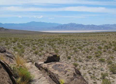 a rock in the foreground serves as a lookout across a desert landscape with a dry lakebed and mountains in the distance
