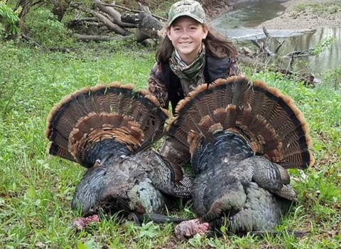 A girl in camouflage crouches on the ground with two large turkeys 