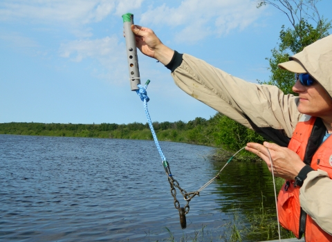 a man holds a temperature logger apparatus - a gray cylinder with a rope and chain attached - at arm's length above the water