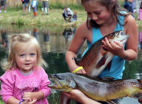 Two girls pose with two big trout in front of a pond where other children are fishing.