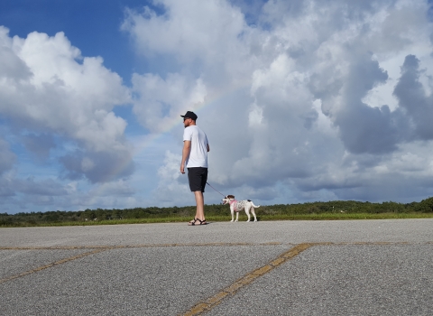 A man walking a dog on a levee with blue skies, white puffy clouds, and a rainbow.