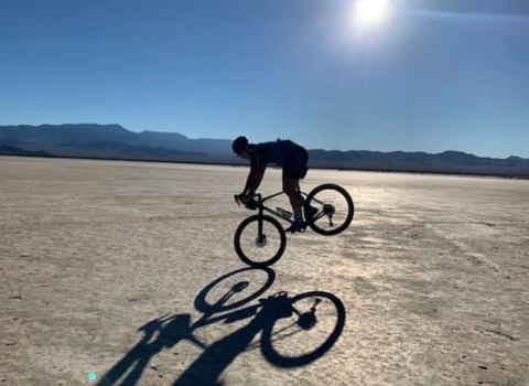 A cyclist's silhouette and shadow are captured on a dry, desert lake bed surrounded by mountains. 