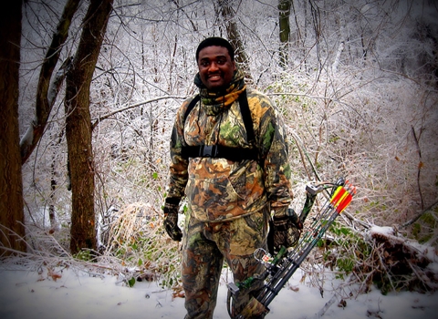 Wayne Hubbard in his hunting fatigue outfit holding his bow and arrow in the woods with snow on the ground. 