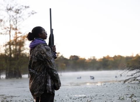 Young Woman In Hunting Gear Stands in wet-marsh at dusk.