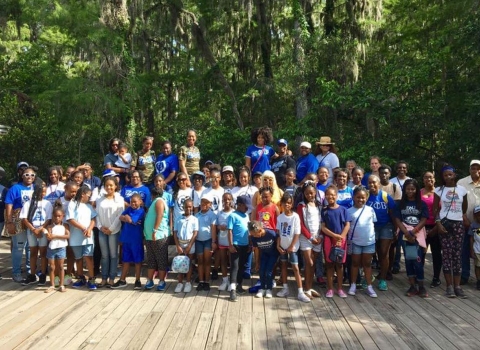 A group of African-American women and children pose for a photo on a boardwalk in front of a forest.