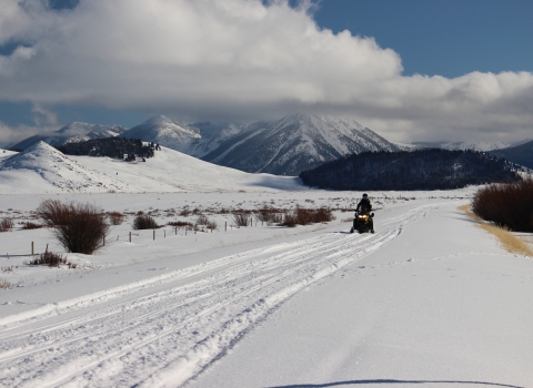 A snowmobiler rides a snowmobile on a snowy road with mountains in the background.