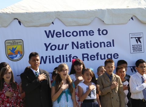 Kids in Sunday clothes with hands over heart in front of sign that says Welcome to Your National Wildlife Refuge