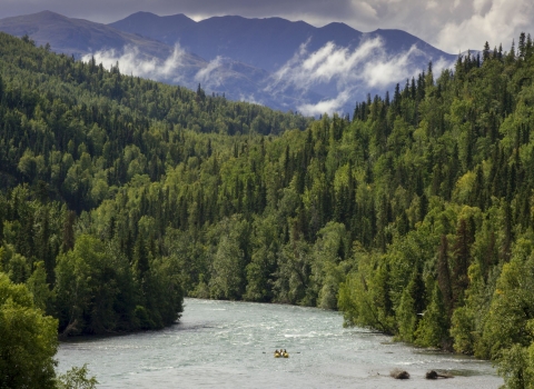A boat heads down a river lined by forests and mountains.
