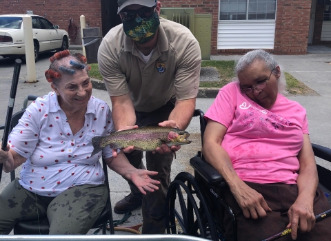USFWS employee holding a Rainbow trout between two seated elderly women