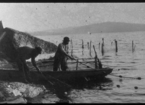 Black and white photo shows two people casting nets from a rowboat