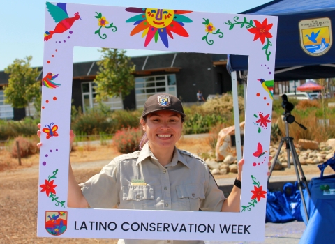 A woman in a brown U.S. Fish and Wildlife Service uniform with a broad smile on her face holding a frame around herself.  On the frame are the words Latino Conservation Week.