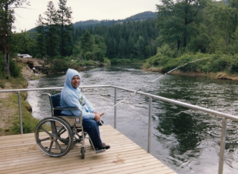 A man in a wheelchair and wearing a hoodie fishing from a platform overhanging a river.
