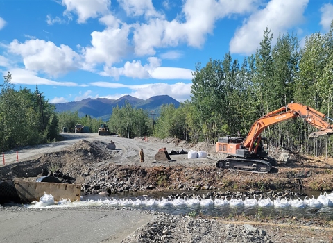 excavator digging near stream with mountains and clouds in background