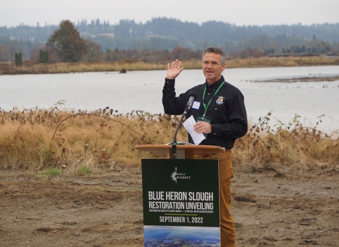 A man in a US Fish & Wildlife Service shirt stands behind a poduum that says "Blue Heron Slough Restoration Unveiling" in front of a body of water