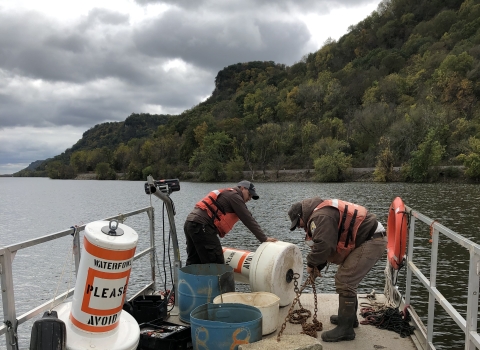 Staff attach a concrete anchor to an orange-and-white Voluntary Waterfowl Avoidance Area buoy before deploying the buoy on the Mississippi River