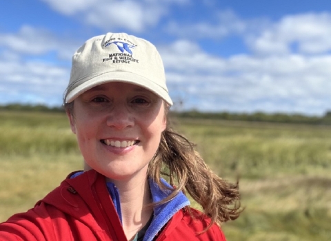 Woman stands in a marsh wearing fish and wildlife hat