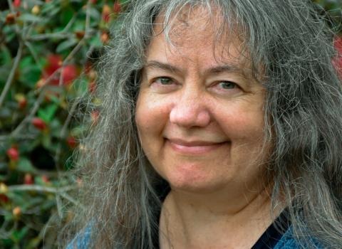 A smiling woman with long, gray hair and blue shirt and sweater stands in front of a green-leaved shrub with orange flowers