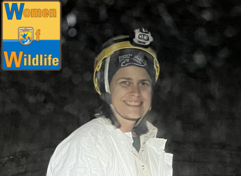 Pam MPam McDill smiles at the camera as she stands in dark cave. She is wearing a white jumpsuit and a helmet with a headlamp on it. The WoW: Women of Wildlife logo is in the upper left corner of the image.