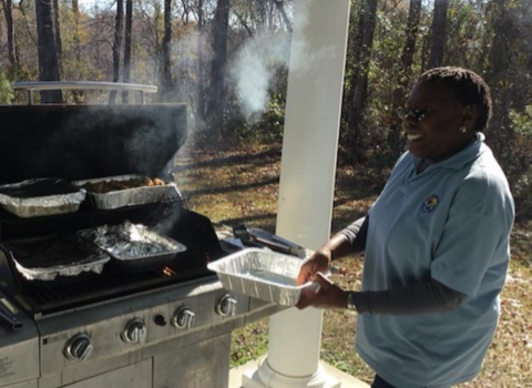 Brenda Williams, volunteer at Waccamaw National Wildlife Refuge, smiles as she holds a tray and stands near a grill where food is being cooked.
