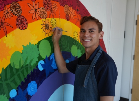 a man smiles while painting a large rainbow mural decorated with various flowers
