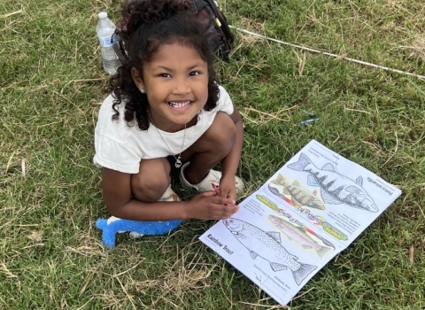 A child looking at the camera smiling and showing off a trout being colored in a coloring book. 