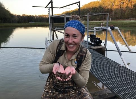Jessica Radich holds mussels while wearing waders and standing in shallow water in front of a dock. 