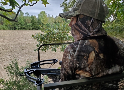 View of a field looking over the left shoulder of a hunter dressed in camouflage with a crossbow in a tree stand.