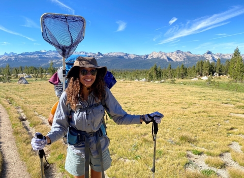 A woman smiles for the camera while holding hiking poles and while wearing a backpack with an attached net on her back