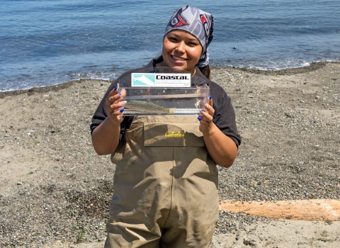 Service Tribal intern standing along a shoreline holding a fish in a small tank.
