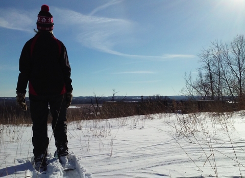 A person wearing snowshoes and a winter hat with a red pompom faces away from the camera as they look out across the Minnesota River Valley from the field next to the overlook behind the Bloomington Education and Visitor Center.