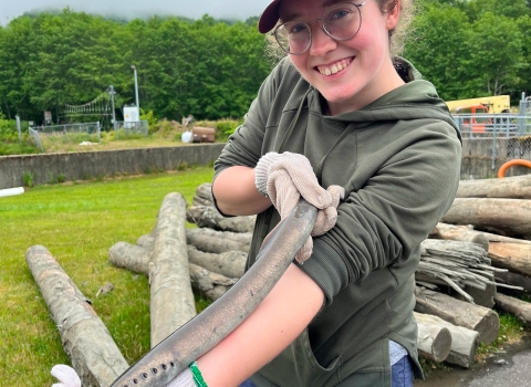 A woman smiles while holding a Pacific lamprey. Logs and structures from Makah National Fish Hatchery are in the background below the trees and sky.