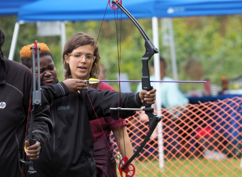 A young adult pulls back on a bow with an arrow drawn, an instructor standing behind him 