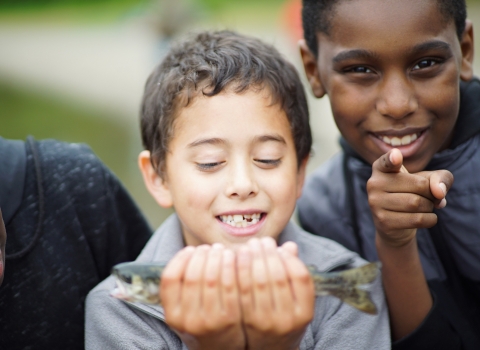 A young child looks at a fish he is holding. A child next to him smiles and points at the camera. 