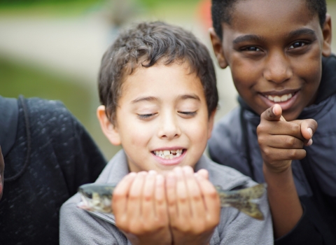 A young child looks at a fish he is holding. A child next to him smiles and points at the camera. 