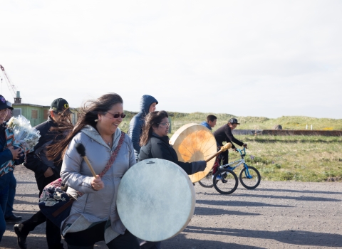 Woman with white  skin drum leads group of people walking down a road. Kids on bikes flank her.