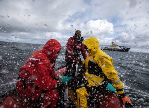 People in bright orange and yellow rain gear in an orange skiff boat through choppy waters through a rain spattered lens. A blue and white ship is in the background.