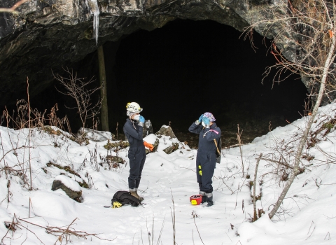 Two people stand in the snow outside a mine entrance, making adjustments to their helmet and face mask