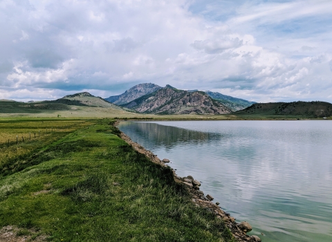 A lake abuts green grass, in the background are small mountains