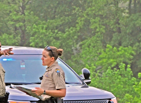 Two law enforcement officers -- a man and a woman -- speaking as the male officer points. They are in front of a police car in a wooded setting.