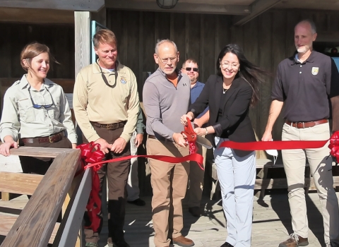 A group of five men and women cutting a ribbon with a pair of oversized scissors. There are a few onlookers in the background.