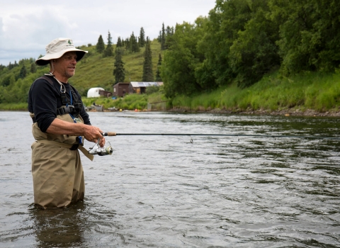 Man in fishing gear boots fishing in a river with hill of green grass and trees.