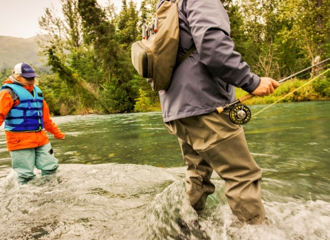 a low-angle shot of a man and a woman in fishing gear standing in rushing river water. The man is holding a fishing pole.
