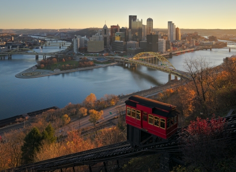 a city skyline photo taken from a hillside. Skyscrapers surrounded by a river, and multiple bridges reaching across the water. A funicular car can be seen in the foreground, as well as trees lining a street. It appears to be autumn due to the vibrantly colored trees.