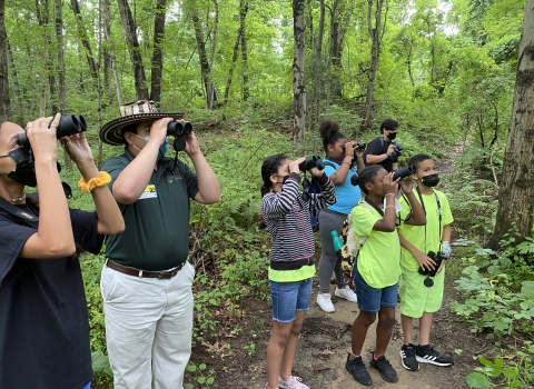 a group of adults and children bird watching with binoculars in a vibrant green forest